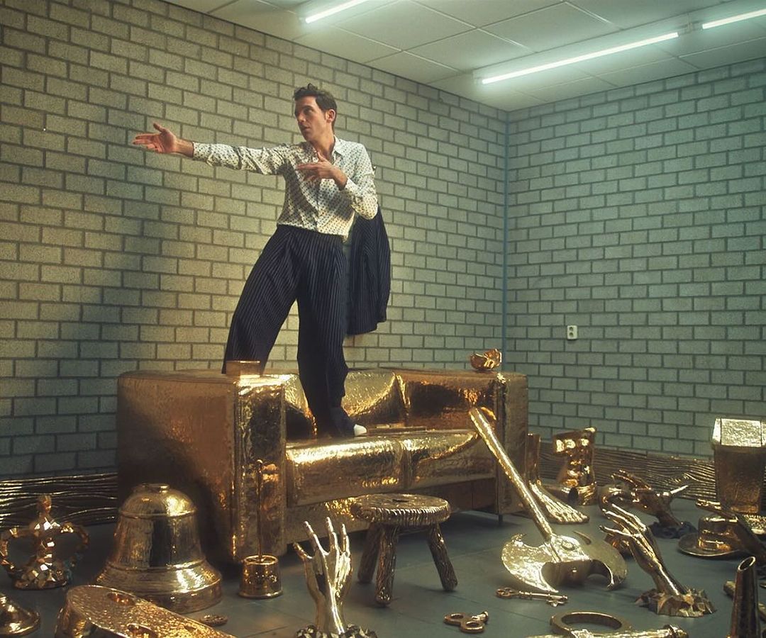 Man stands on a gold couch surrounded by gold objects in a brick-walled room under fluorescent lighting, casting shadows and highlighting textures.