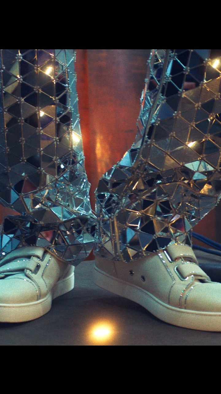 Close-up of lower body wearing mirror-shard pants and white sneakers. Abstract reflective geometric pattern covers pants and shoes.