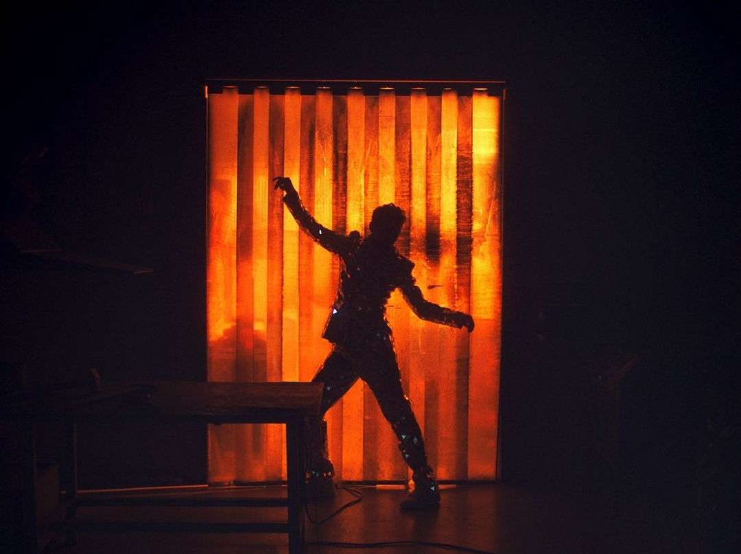 Silhouette of a performer in a sequined outfit against a bright orange and yellow backdrop during a concert performance.
