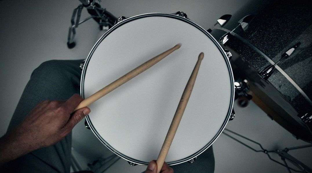 Overhead view of drum with drumsticks held by a person wearing grey sweatpants in a music setting under studio lighting.