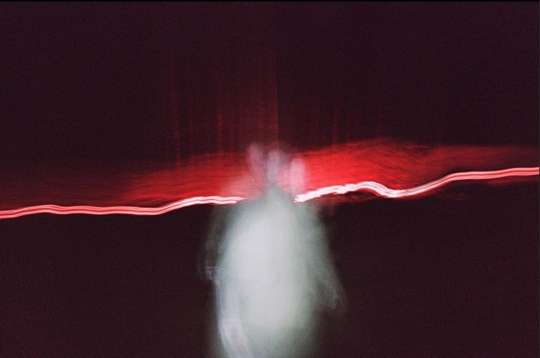 Abstract long exposure shot with blurred figure, red light streaks, and dark background, creating a mysterious and ethereal aesthetic.