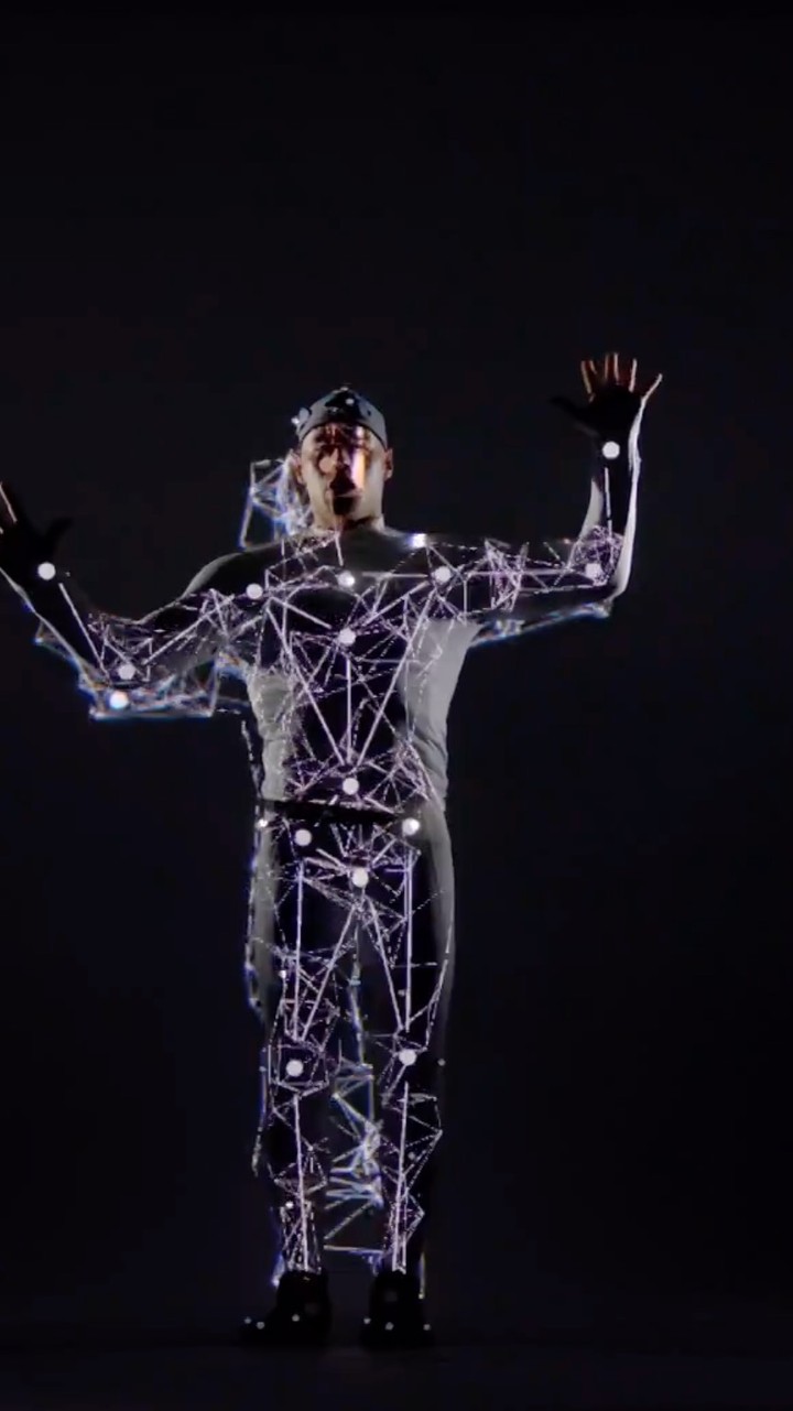 Man in a motion capture suit stands against a dark background with arms raised, covered in a pattern of white dots and lines.