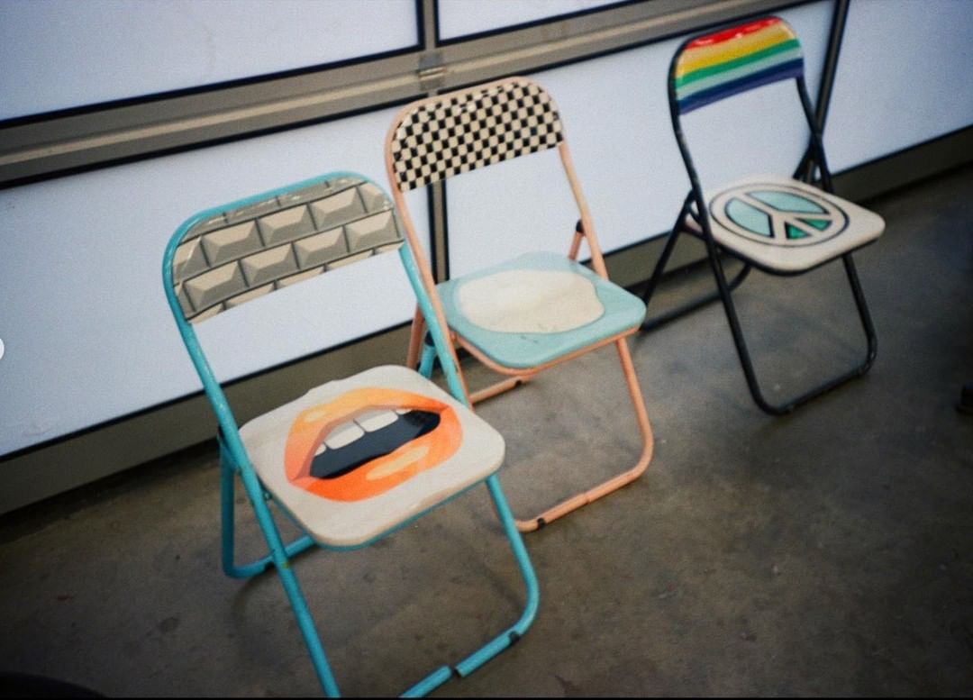 Three folding chairs with playful designs, including lips, a checkerboard pattern, and a peace sign, stand indoors against a wall.