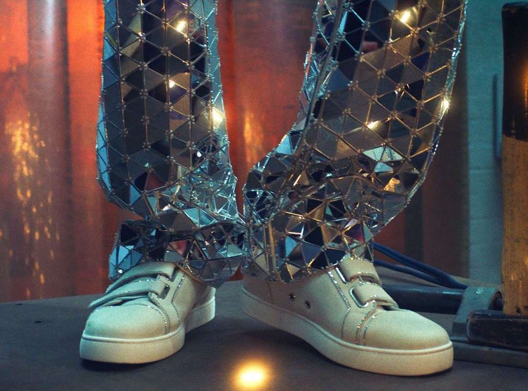 Person wearing reflective mosaic-tiled pants and white sneakers stands indoors. The pants feature a grid of triangular mirrors.
