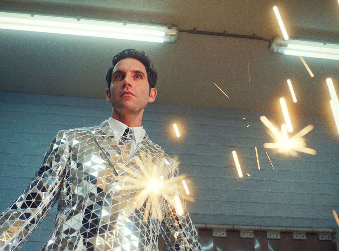 Adult male in disco ball suit looking up and to the left with light flares, indoor in front of brick wall with light fixture.