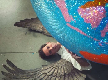 Man lying on a grey floor wearing angel wings and a white shirt holding a large sparkling globe