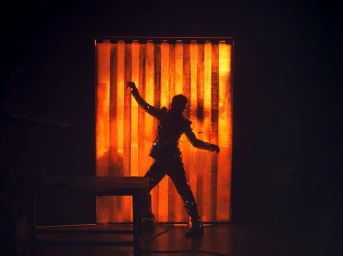 Silhouette of a dancing figure posed in front of a vertically striped glowing orange light panel in a dark room.