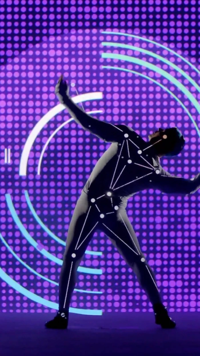 A dancer wearing a motion capture suit with glowing tracking nodes poses against a vibrant purple dotted digital background screen.