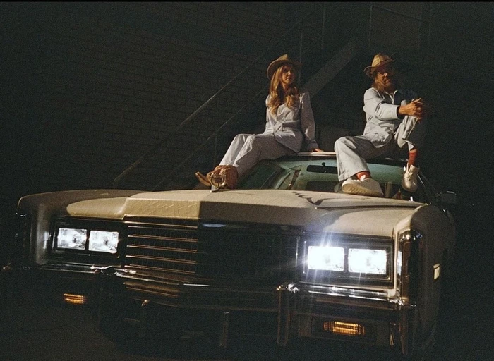 Man and woman wearing matching light-colored suits and hats seated on the hood of a vintage white car with headlights on at night.