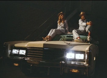 Man and woman wearing matching light-colored suits and hats seated on the hood of a vintage white car with headlights on at night.
