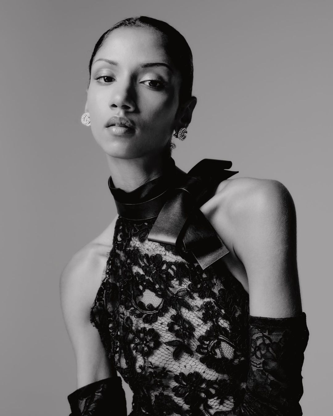 Black and white portrait of a woman in a lace dress with a high collar and bow detail, accessorized with earrings and gloves.