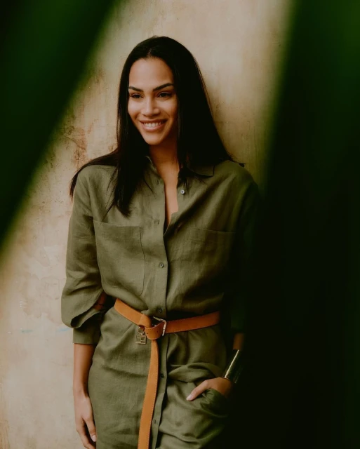 Smiling woman in an olive green jumpsuit with a tan belt, standing indoors with soft lighting and green plant out of focus.