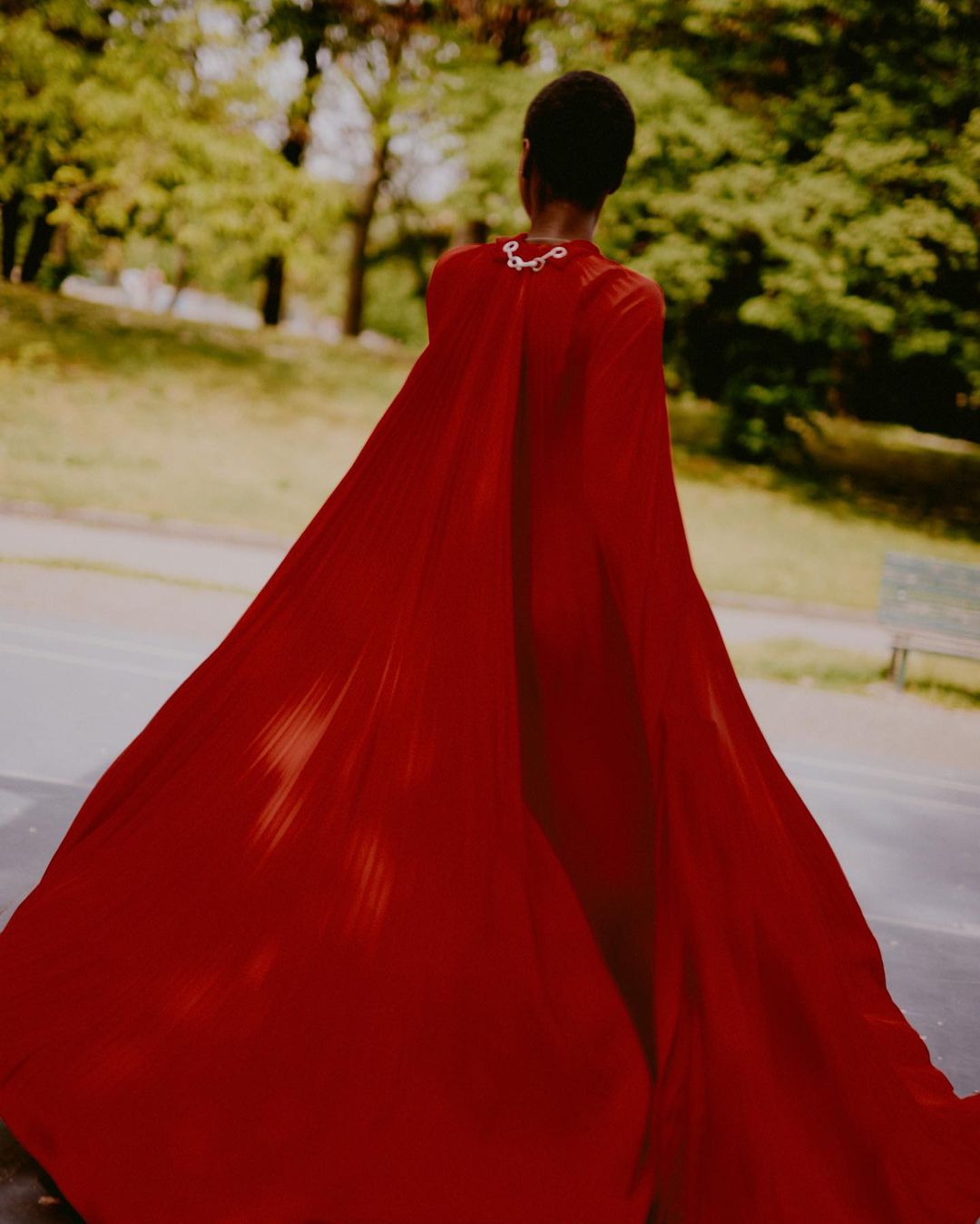 Woman in a flowing red gown is walking through a park, captured from behind with blurred trees and greenery visible in the background.