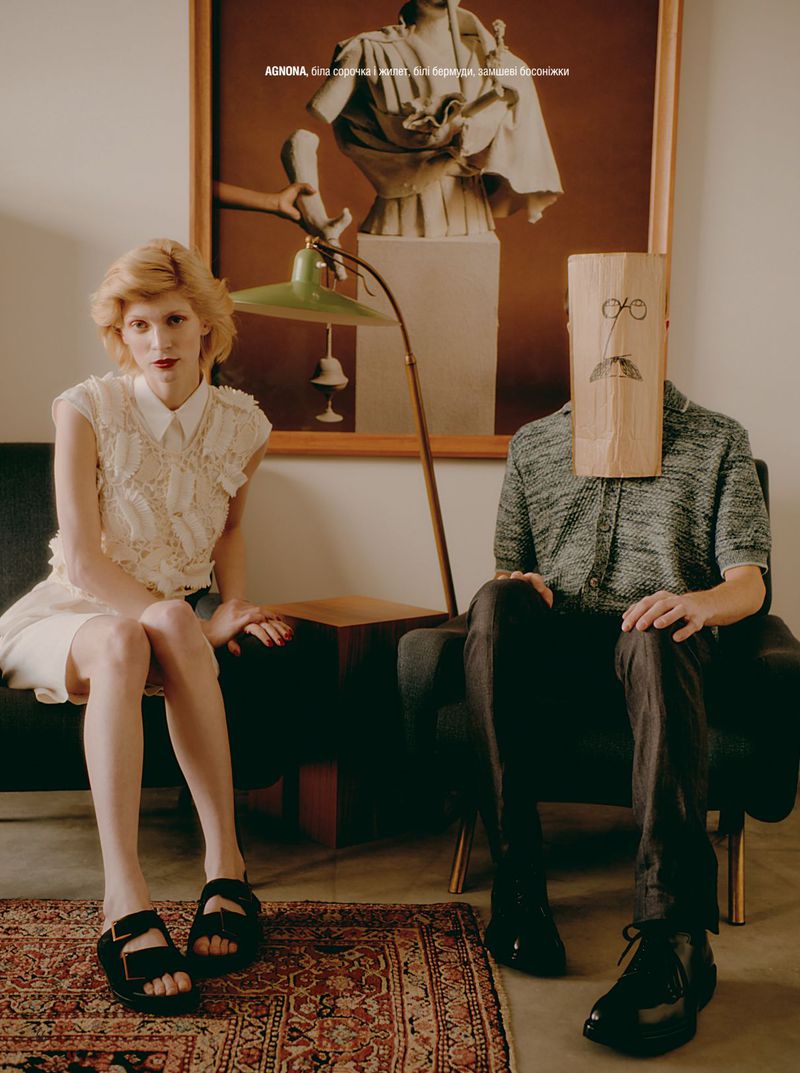 Indoor fashion shot featuring a woman in a white dress and a man with a paper bag on his head in a living room setting.