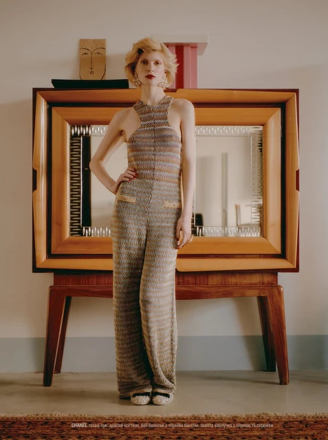 Woman in knitted halter top and pants poses in front of a wooden cabinet indoors. Fashion editorial photography with a retro aesthetic.