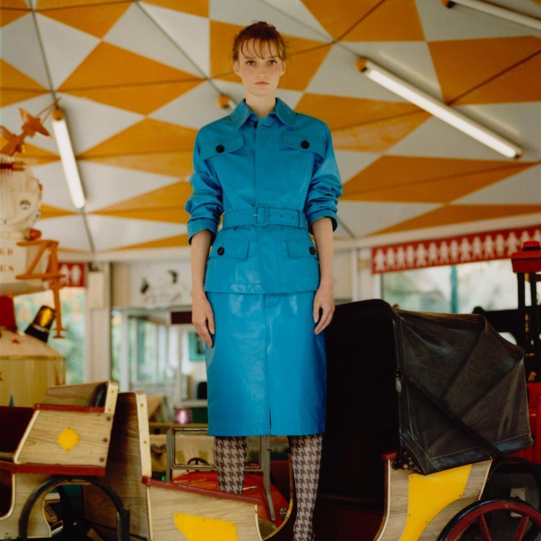 A stylish adult female model poses in an aqua-blue jacket and skirt in a carousel setting, showcasing fashion and editorial style.