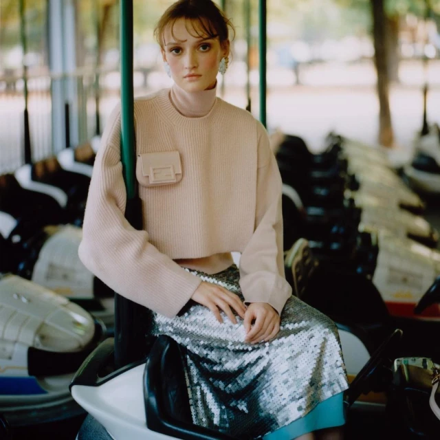 A stylish young woman in a peach sweater and sequin skirt sits on a vintage ride, capturing a blend of fashion and whimsical nostalgia.