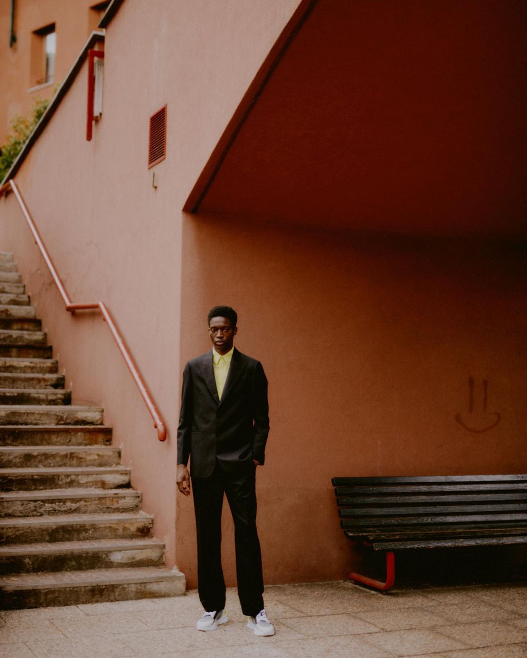Full shot of an adult man in a suit standing outdoors against a terracotta colored building with stairs and a bench.