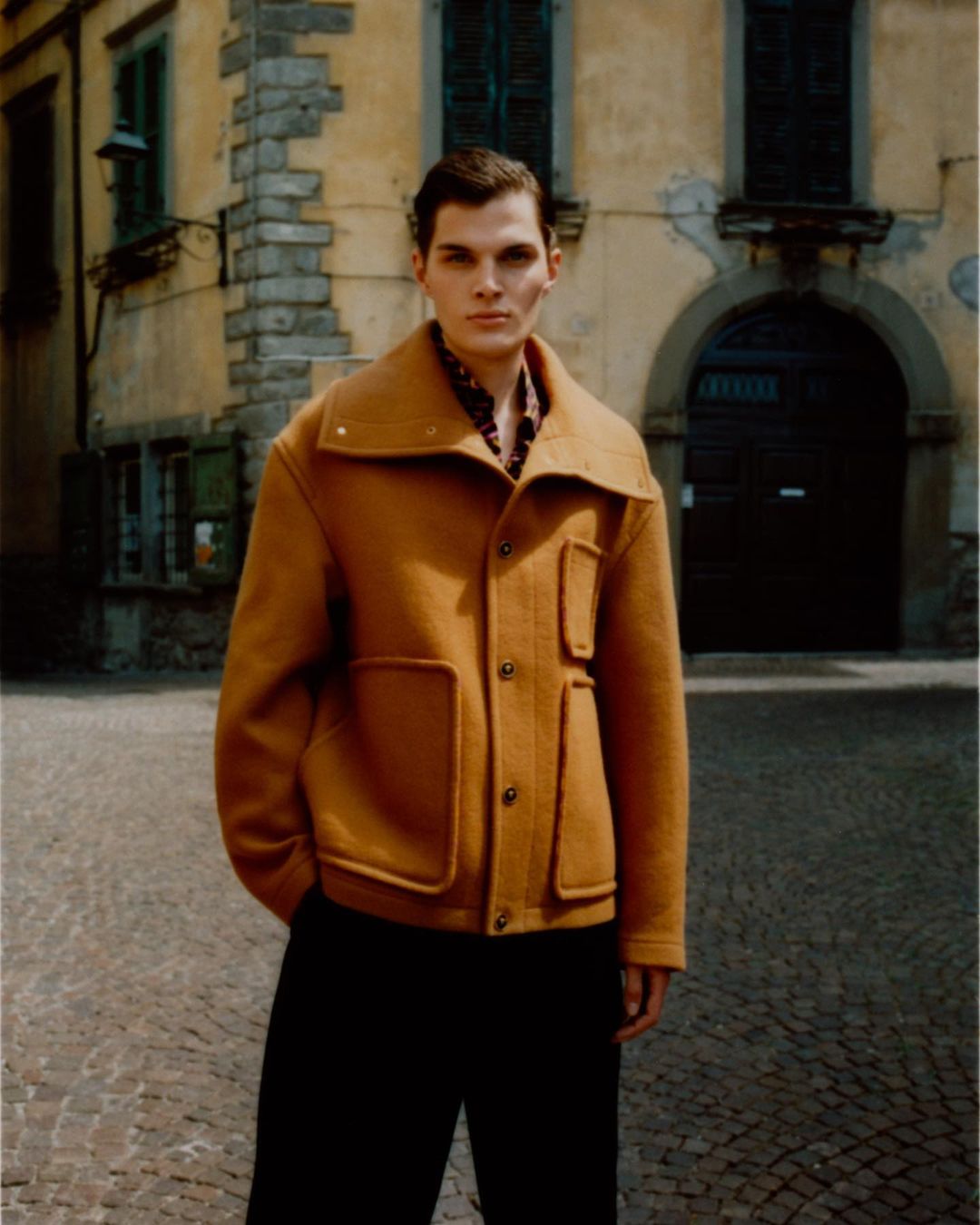 A stylish adult male stands outdoors wearing a gold jacket and black pants against an old European-style building and cobblestone pavement.