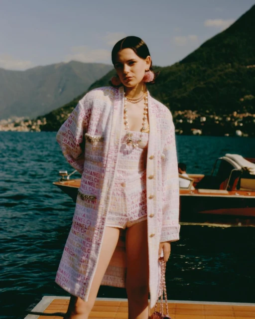 A young woman in a Chanel-branded white short set and matching jacket poses on a wooden dock, with a lake and mountains behind her.