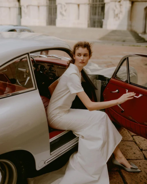 A woman in white dress sits in a silver classic car with a red interior in front of a light stone building on a brick street.