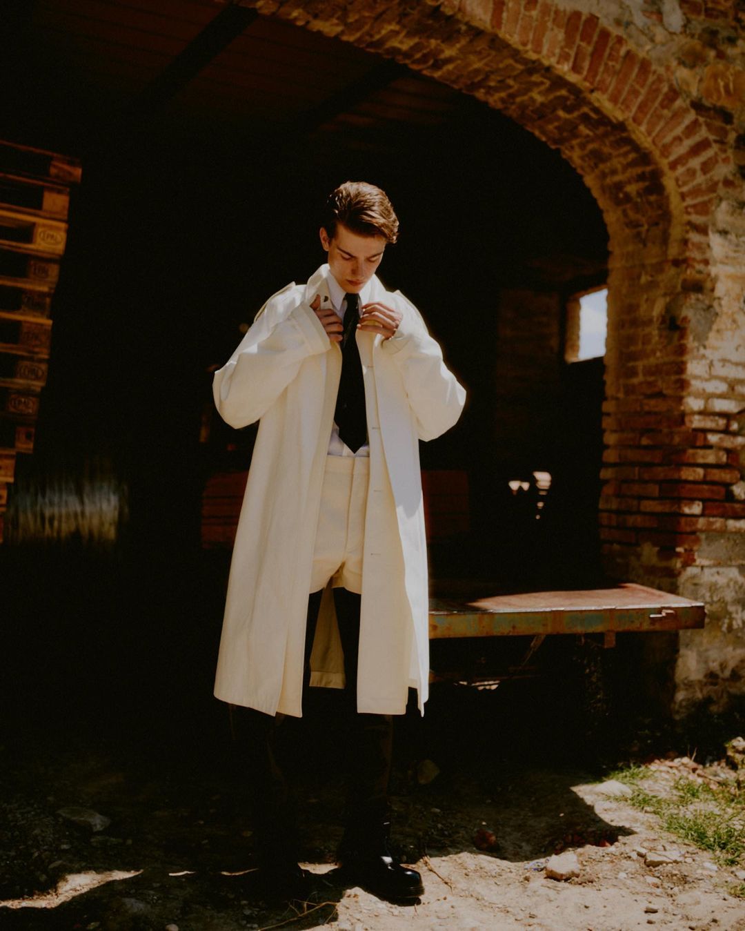 Young man in an off-white coat and suit standing indoors in front of an arched doorway and wearing a dark tie and black boots.