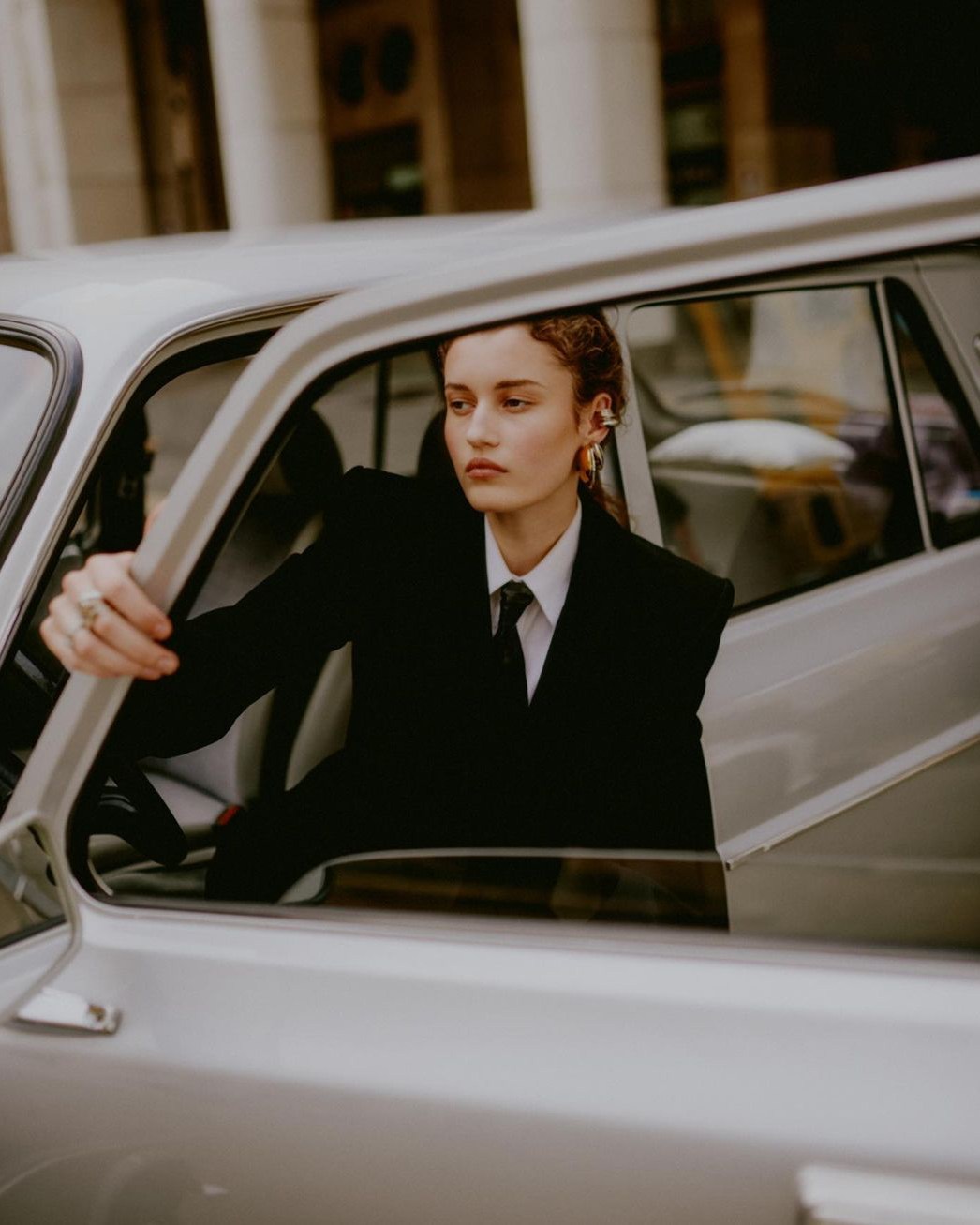 Woman in a black suit jacket and white shirt sitting inside a vintage silver car, photographed by Federico Barbieri in Milan.