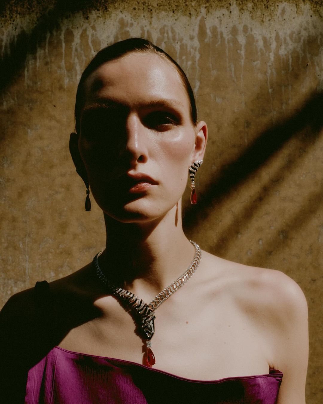 A model poses in an indoor fashion shot with bright magenta top, wearing statement necklace and earrings in front of textured wall.
