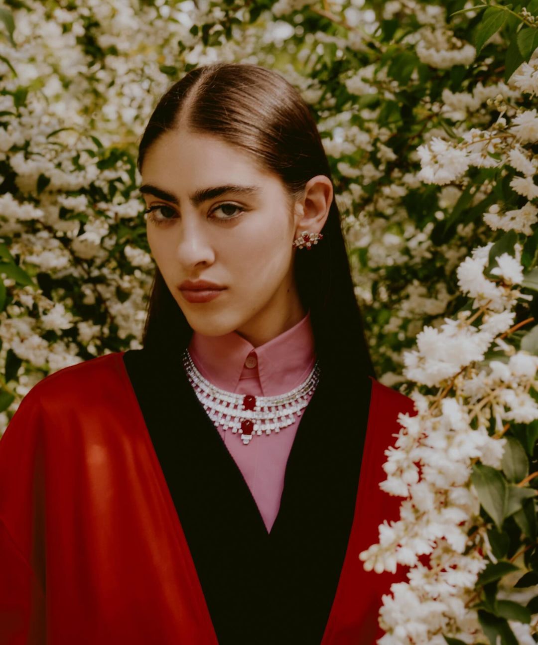 A young woman poses in a red cape and pink shirt with a layered silver and red beaded necklace, wearing matching earrings outdoors.