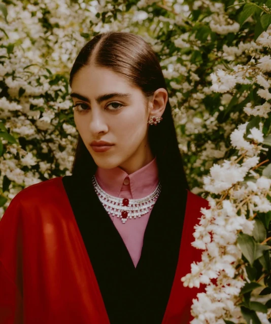 A young woman poses in a red cape and pink shirt with a layered silver and red beaded necklace, wearing matching earrings outdoors.