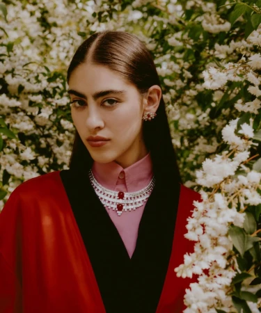 A young woman poses in a red cape and pink shirt with a layered silver and red beaded necklace, wearing matching earrings outdoors.