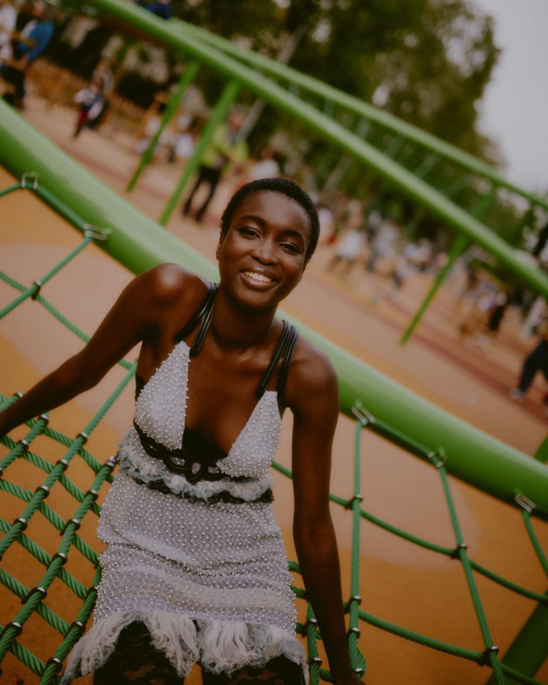 A smiling African woman in a light dress poses on a green structure at a playground with blurred people in the background.