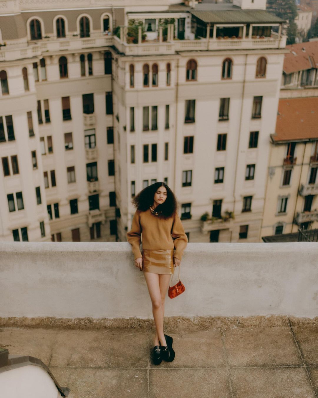 Woman with dark curly hair wearing a brown sweater and mini skirt standing on a rooftop in Milan by Federico Barbieri