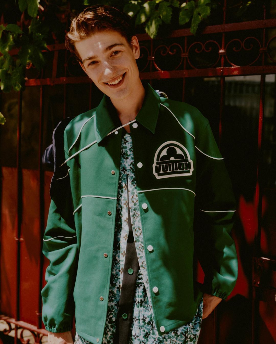 Smiling adult male in a green jacket with a floral shirt, standing outdoors with a red gate and green foliage visible.