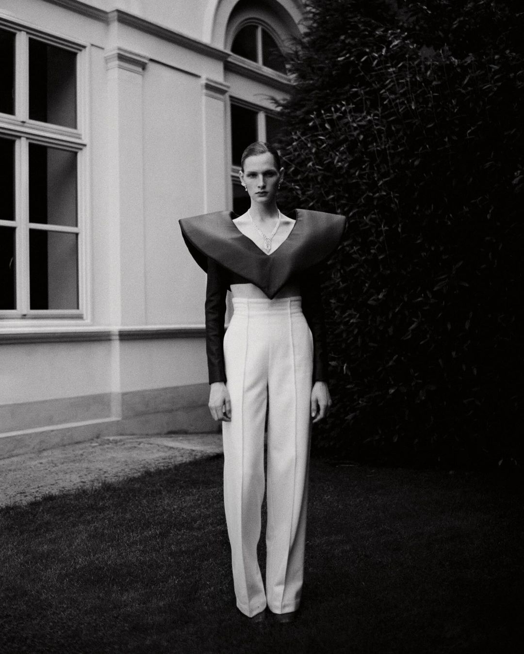 Monochrome fashion portrait of a slender adult with high-waisted white pants and a bold, oversized burgundy collar piece in a grassy outdoor setting.