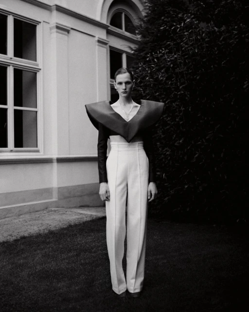 Monochrome fashion portrait of a slender adult with high-waisted white pants and a bold, oversized burgundy collar piece in a grassy outdoor setting.