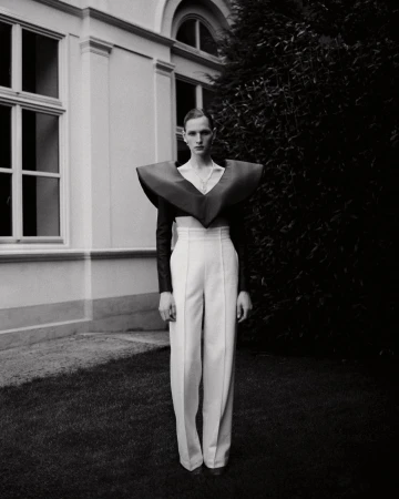 Monochrome fashion portrait of a slender adult with high-waisted white pants and a bold, oversized burgundy collar piece in a grassy outdoor setting.