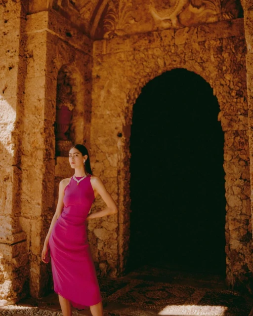 Young woman in elegant fuchsia dress poses in stone archway with decorative carvings; fashion shoot in historic setting.