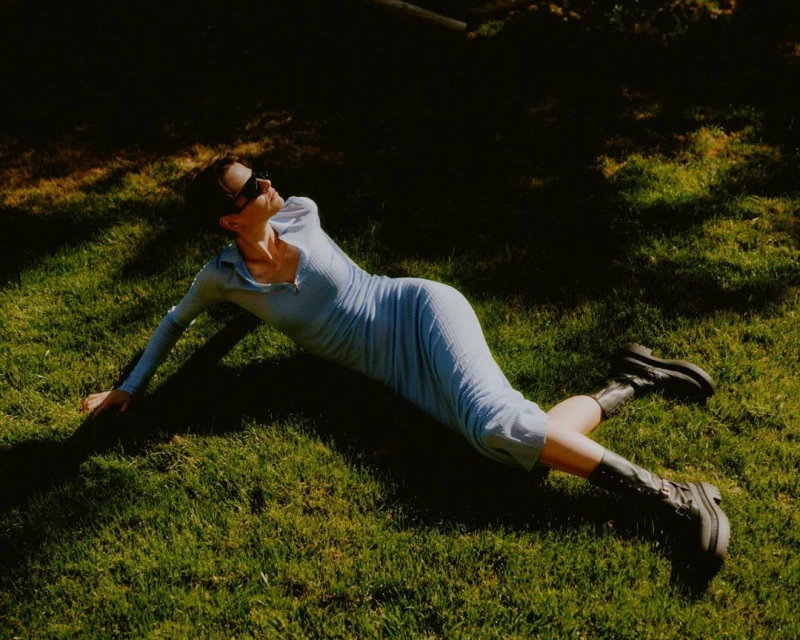 Woman in a blue dress and boots lies on green grass in sunlight, enjoying a serene outdoor moment, capturing a blend of fashion and nature.