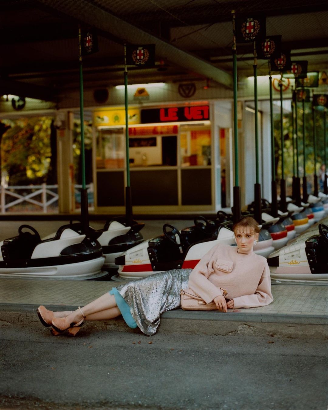 Woman in a light pink sweater and silver skirt lies on a concrete ledge in front of a row of bumper cars in an amusement park.