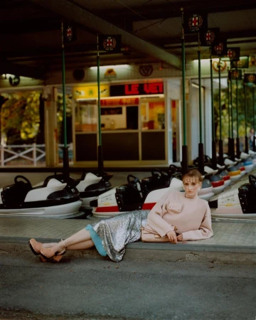 Woman in a light pink sweater and silver skirt lies on a concrete ledge in front of a row of bumper cars in an amusement park.