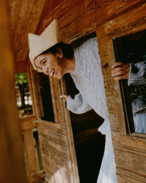 Woman smiling and looking through the window of a wooden playhouse. The woman is wearing a light blue sweater and a paper hat.