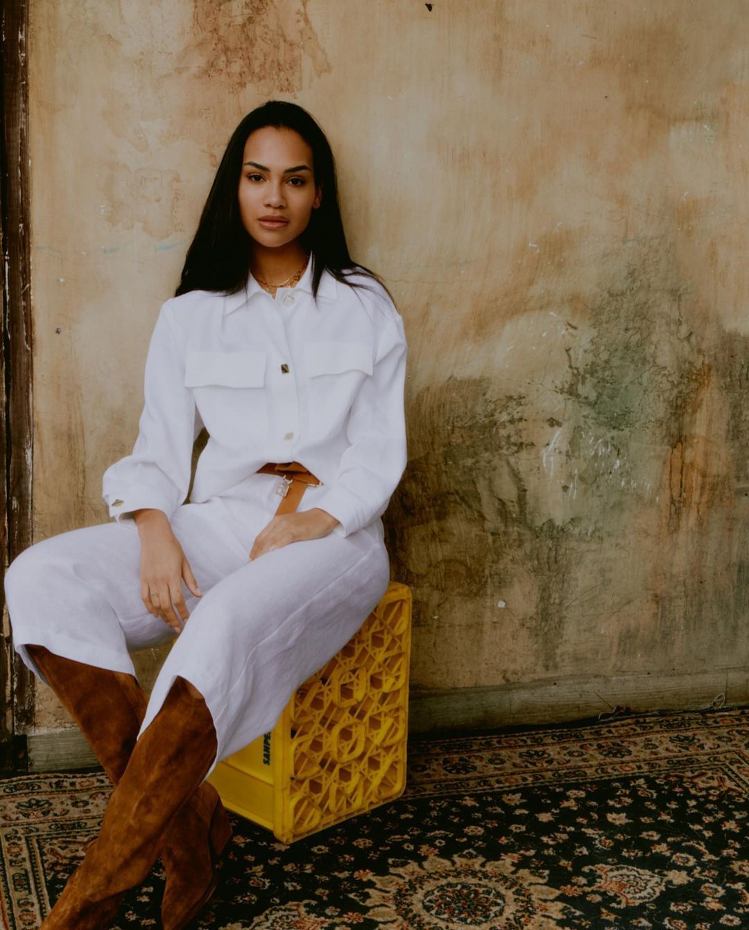 A woman in a white linen outfit poses in front of a mottled wall while seated on a yellow crate, wearing brown boots on a patterned rug.