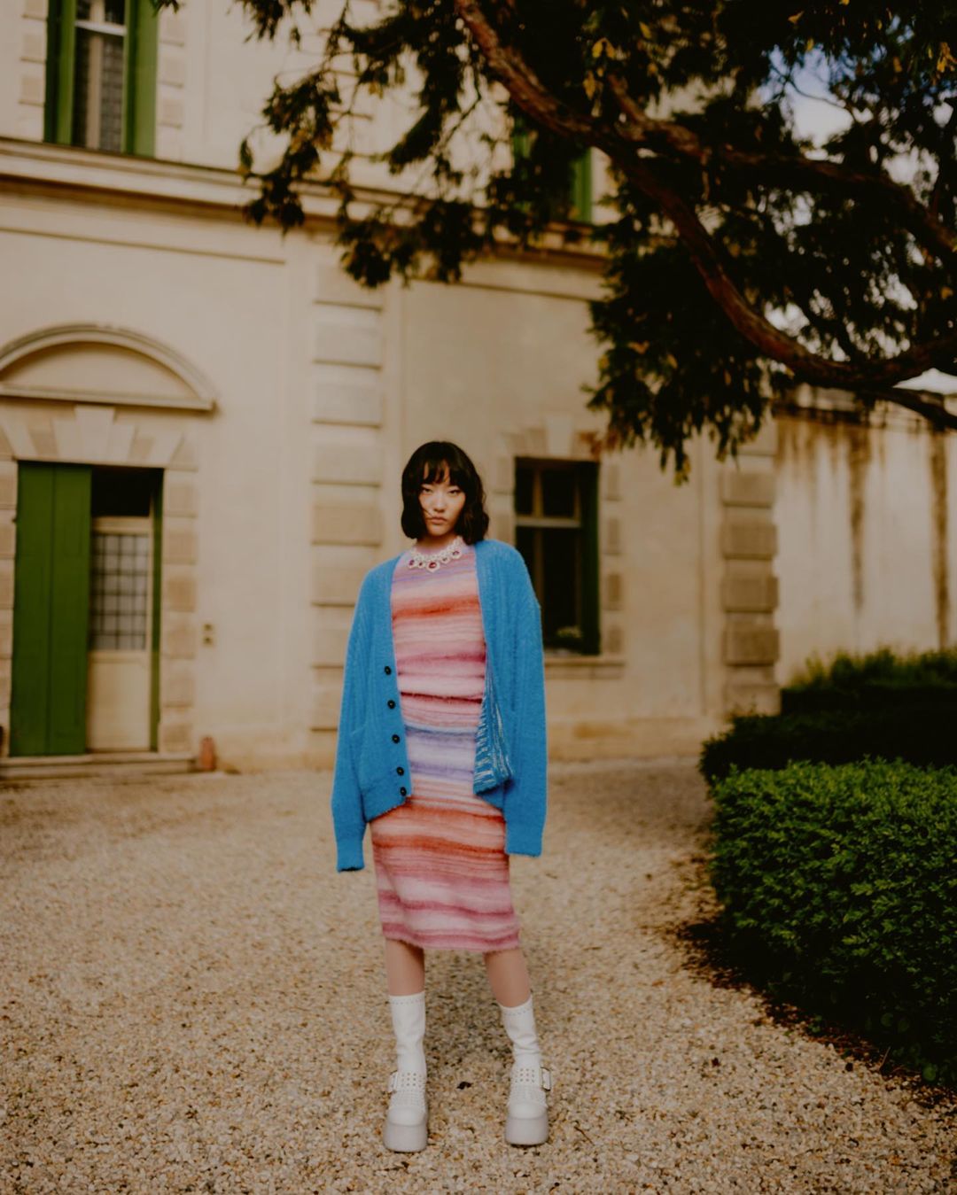 A young Asian woman poses in a colorful knit dress and blue cardigan, set against a tan building with green shutters and an old tree.
