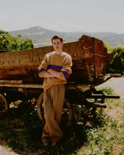Young adult man in neutral sweater with colored stripes poses outdoors, leaning against an old wooden cart with mountain background.