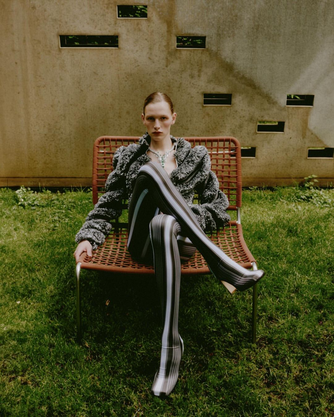 A fair-skinned adult woman sits in a wicker chair in a grassy outdoor setting wearing a patterned jacket and striped stockings.