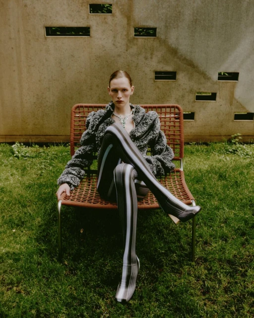 A fair-skinned adult woman sits in a wicker chair in a grassy outdoor setting wearing a patterned jacket and striped stockings.