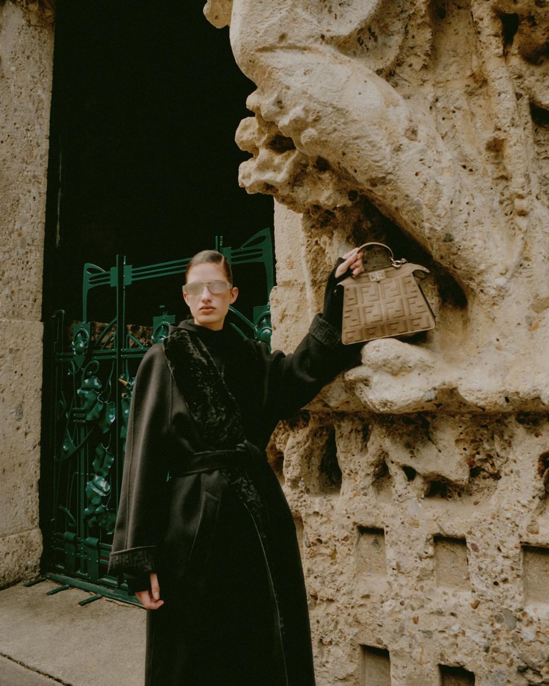 Woman in a long black coat holds a Fendi purse next to a stone wall and a dark green decorative iron gate; outdoor fashion.