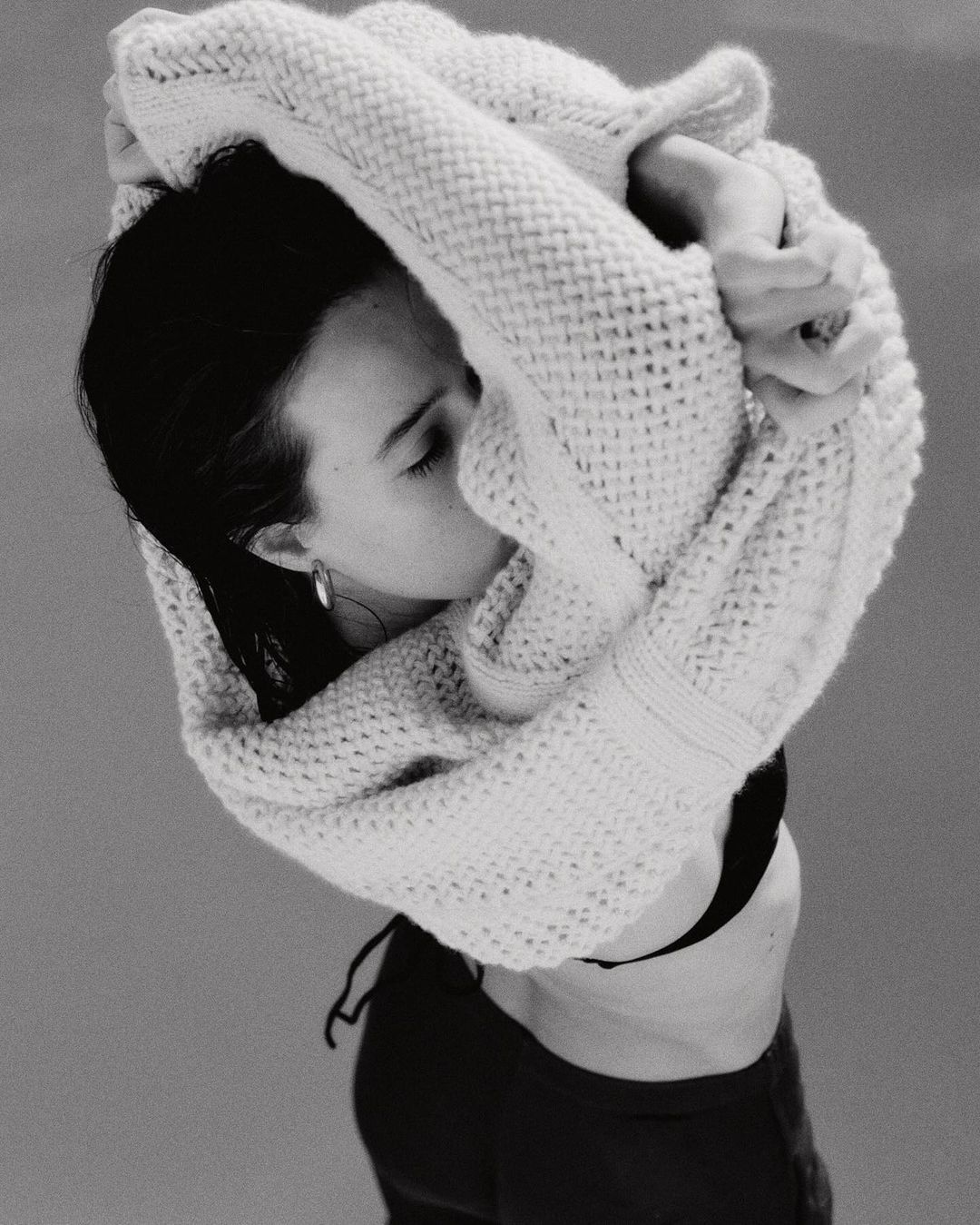 Monochrome portrait of a woman in a black top and cardigan, gazing down. The shot is from above with soft lighting and minimal background.