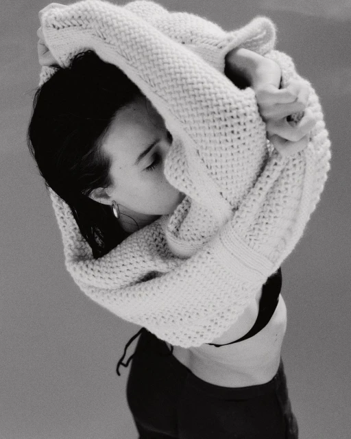 Monochrome portrait of a woman in a black top and cardigan, gazing down. The shot is from above with soft lighting and minimal background.